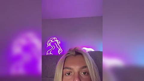 Media: A video of a blonde woman with light skin, wearing a gray sweater, with a neon light of a woman in a dress illuminating the background.