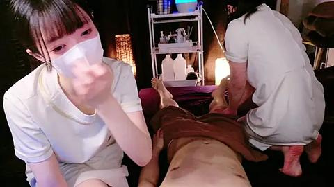 Media: Video of two Asian masseuses, one in a white t-shirt, the other in a white shirt and pants, massaging a nude Asian man on a massage table in a dimly lit room.