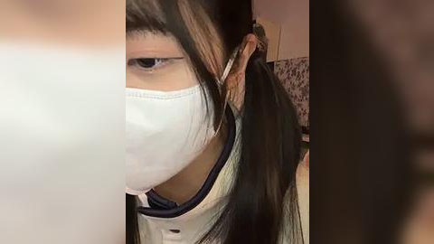 Media: Video of an Asian woman with long black hair, wearing a white face mask and a black and white striped shirt, seen from the side, in a dimly lit room with blurred background.