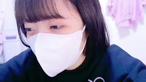 Media: Video of an Asian woman with straight, dark hair and bangs, wearing a white surgical mask, black top, and purple shirt. Background shows a white room with hanging clothes.