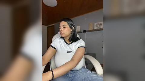 Media: Video of a young woman with medium skin tone, dark hair, and a white t-shirt with black trim, sitting on a white office chair, appearing tired, in a dimly lit room with wooden ceiling, shelves, and a closed door.