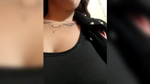 Media: A close-up video of a woman with fair skin, wearing a black top and black gloves, revealing a detailed, black ink tattoo of a mandala on her chest.