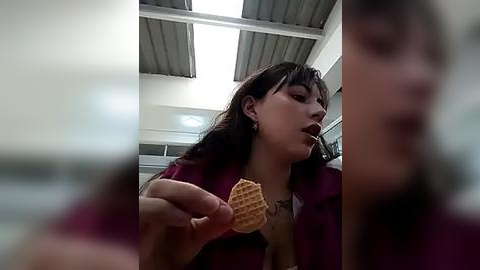 Media: A video of a young woman with long black hair and bangs, wearing a magenta jacket, holding a waffle in a modern, brightly lit room with white walls and a metallic ceiling.