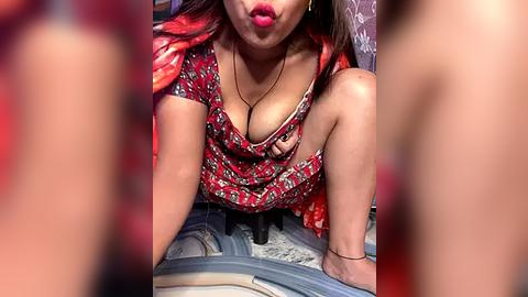 Media: Video of a young woman with medium skin tone and long dark hair, wearing a low-cut red dress with floral print, sitting on a car seat, pouting with red lipstick.