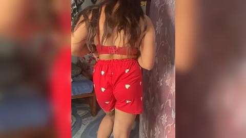 Media: Video of a woman with long, wavy brown hair in red pajama shorts and a matching bra, leaning against a floral-patterned curtain. Background shows a bedroom with a bed and scattered objects.