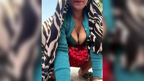 Media: Video of a woman with medium skin tone, wearing a turquoise cardigan exposing cleavage, red patterned skirt, and black zebra-print scarf, kneeling outdoors.