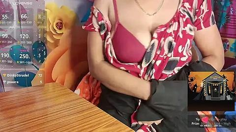 Media: A video of a woman in a red floral top, revealing a pink bra, sitting at a wooden table. Background features a colorful, abstract wall painting. TV screen displays weather and news.