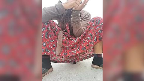 Media: Video of a person squatting in a red skirt with a pattern, wearing a brown top and black socks with brown shoes, against a plain white background.