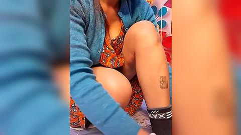 Media: Video of a person in a blue cardigan, orange floral dress, and black socks, with a tattoo on the right thigh, sitting on a red background, blurry foreground.