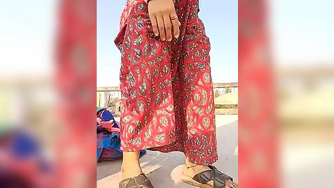 Media: Video of a person wearing vibrant red pants with white floral patterns, standing on a sunny outdoor terrace, with a blurred background of a blue sky and a colorful bag.