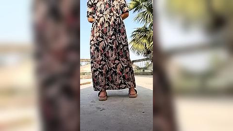 Media: Video of a woman wearing a long, black and white floral dress with brown sandals, standing on a paved path with a blurred background of palm trees and a clear sky.