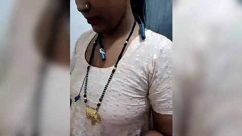 Media: Video of a woman with medium skin tone, wearing a white, floral-patterned blouse and a gold star pendant necklace. Her nose is pierced, and she has a blue bead necklace. Background is blurred, focusing on her face and upper torso.