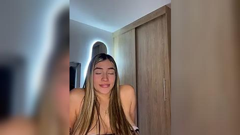 Media: Video of a smiling young woman with long, straight brown hair, topless, in a bathroom with wooden closet doors and a mirror.