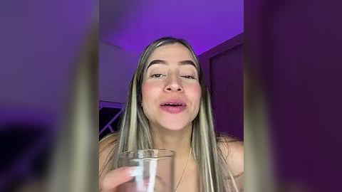 Media: Video of a young Caucasian woman with long, straight blonde hair, fair skin, and light makeup, holding a glass of water. Background shows a dimly lit room with purple lighting.