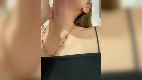 Media: A video of a fair-skinned woman with blonde hair, wearing a black spaghetti-strap top, and a gold chain necklace, partially obscured by a blurred foreground object.