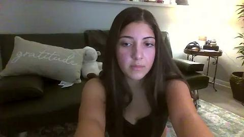 Media: Video of a young woman with long dark hair, fair skin, and a neutral expression, sitting on a green leather couch in a living room.