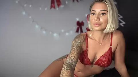 Media: Video of a blonde, fair-skinned woman with tattoos, wearing red lace lingerie, posing seductively against a festive, softly lit background with white string lights and red bows.