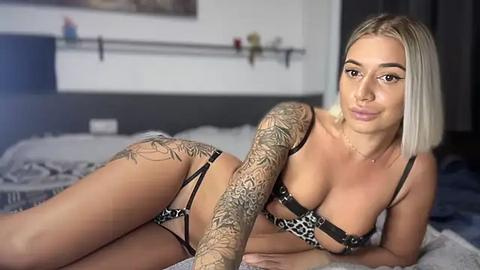 Media: Video of a blonde woman with medium breasts, wearing leopard print lingerie, lying on a bed with intricate arm tattoos, in a modern bedroom.