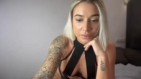 Media: Video of a blonde woman with light skin and tattoos, wearing a black halter top, resting her chin on her hand, looking thoughtful.