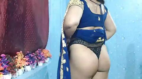 Media: Video of a dark-skinned woman in a blue and gold sari, showcasing her curvy buttocks in black lace panties, against a light blue wall with floral decorations.