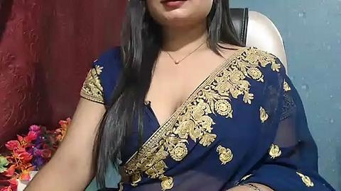 Media: Video of a woman with long black hair, wearing a blue sari with gold embroidery, seated in a chair. Background includes red curtains and a floral arrangement.