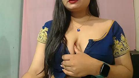 Media: Video of a South Asian woman with long black hair, wearing a blue blouse with gold embroidery, exposing her ample cleavage. She has a gold necklace and a black smartwatch on her wrist. Pink and green walls in the background.