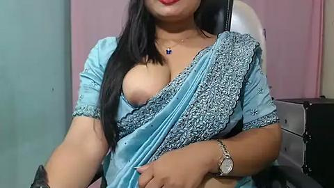 Media: Video of an Indian woman with medium skin tone, wearing a partially open blue sari, revealing one breast. She sits in a chair with a pink wall and a silver object in the background.