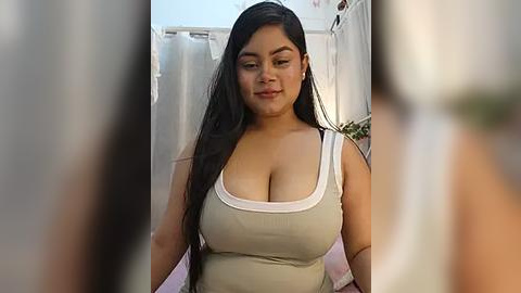 Media: Video of a curvy Latina woman with long black hair and large breasts, wearing a beige tank top. She stands in a softly lit, blurry bathroom with white walls and a shower curtain.