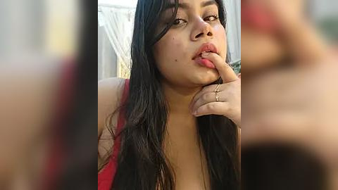 Media: Video of a young woman with long black hair, fair skin, and full lips, wearing a red top, sensually biting her index finger, blurred background.