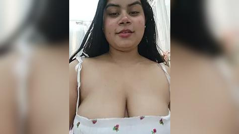 Media: Video of a smiling, light-skinned woman with long black hair, wearing a white spaghetti-strap top with floral print, her large breasts prominently displayed.