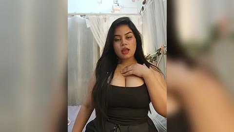 Media: A video of a curvy, light-skinned woman with long black hair, wearing a black top, standing in a garden gazebo. Her left hand touches her chest, emphasizing her ample cleavage.