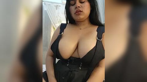 Media: Video of a voluptuous, light-skinned woman with long black hair, wearing a revealing black corset dress with low-cut neckline, exposing ample cleavage. Background features blurred, neutral-colored walls.