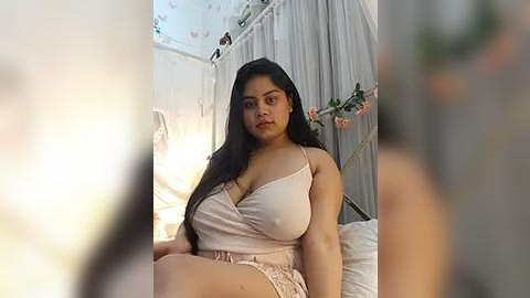 Media: A video of a plus-sized Latina woman with long black hair, wearing a pink spaghetti-strap top and shorts, sitting on a bed with a white canopy, surrounded by floral decorations.