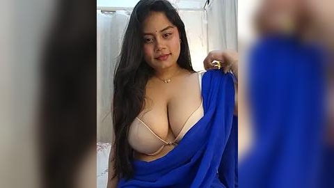 Media: Video of a curvy South Asian woman with long black hair, holding a blue sari, wearing a beige bra, and a necklace, indoors, blurred background.