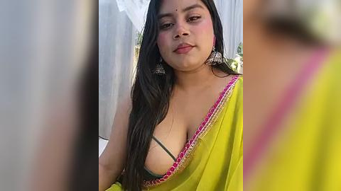 Media: Video of a young South Asian woman with long black hair, wearing a yellow sari with pink trim, exposing a green bra, standing against a white curtain backdrop.