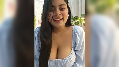 Media: Video of a smiling Latina woman with long black hair, wearing a light blue top that accentuates her large breasts, standing outdoors with blurred background and greenery.