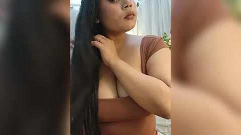 Media: A video of a plus-sized woman with medium skin tone, long black hair, and a brown top, touching her neck, set against a blurred background.