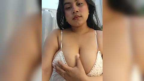 Media: Video of a plus-sized woman with medium brown skin, dark hair, wearing a lacy white bra, touching her large breasts, in a bathroom with towels hanging.