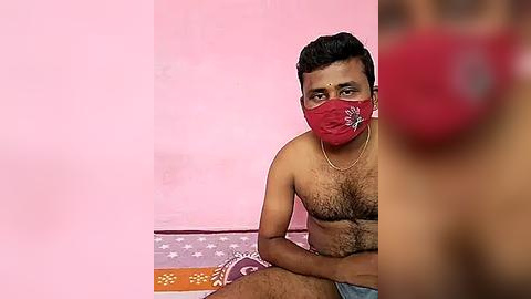 Media: Video of a South Asian man with short black hair, wearing a red mask, red and white printed T-shirt, and blue shorts, sitting against a pink wall.