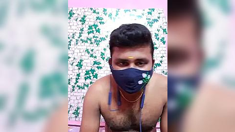 Media: Video of a shirtless, muscular South Asian man with short black hair, wearing a blue mask and gold necklace, against a floral-patterned wall.