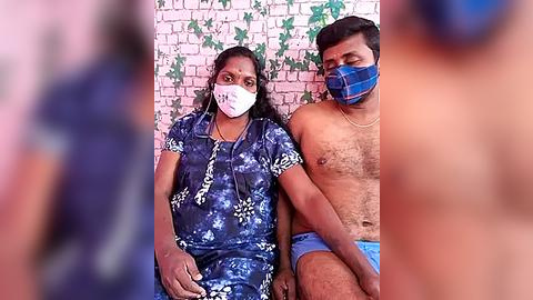 Media: Video of a South Asian woman in a blue floral dress, wearing a face mask, sitting beside a shirtless South Asian man in blue shorts, both against a white brick wall with green ivy.