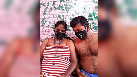 Media: Video of a pregnant woman with short dark hair, wearing a striped top and black face mask, sitting next to a shirtless man with a beard, both against a white brick wall with green ivy.