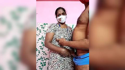 Media: Video of an Indian woman in a floral dress with a white mask, checking the size of a man's blue boxer shorts, in a tiled room with green and white patterned walls.