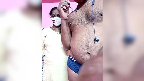 Media: Video of an overweight man in blue boxers and a stethoscope, standing next to a slim woman in a yellow dress and face mask, in a brightly lit room with pink walls.