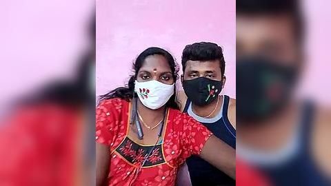 Media: Video of two South Asian people with dark skin, wearing face masks and traditional red outfits, standing against a pink wall, blurred background.