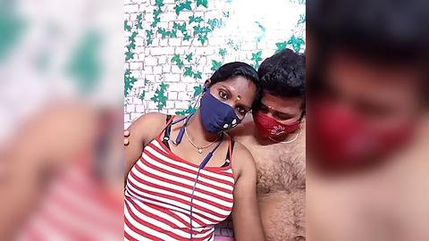 Media: Video of a young woman with medium brown skin, wearing a red-and-white striped tank top and blue face mask, hugging a shirtless man with medium brown skin, wearing a red face mask, both leaning against a white brick wall with green vines.