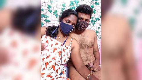 Media: Video of a South Asian couple indoors, wearing face masks; man is shirtless, woman in floral dress, with a white brick wall and green leaf patterned background.