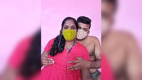 Media: Video of an Indian woman wearing a bright pink saree, yellow mask, and a bindi, standing with a shirtless man with dark skin, wearing a white mask, in a pink room.