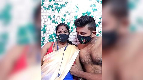 Media: Video of a South Asian woman and man, wearing masks, against a white brick wall with green ivy. The man, shirtless, is muscular, while the woman wears a red sari.