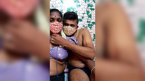 Media: Video of two South Asian women, one with a purple bra and mask, the other topless with a beige mask, embracing in a dimly lit room with green walls.